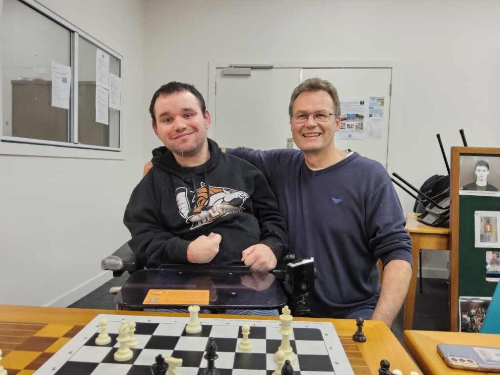 Jono taking a photo with a fellow chess player, in front of a chess board