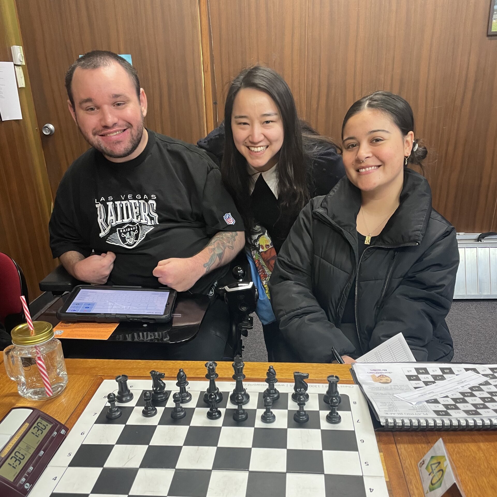 Jono posing for a photo with his friend and a chess arbiter, in front of a chess board