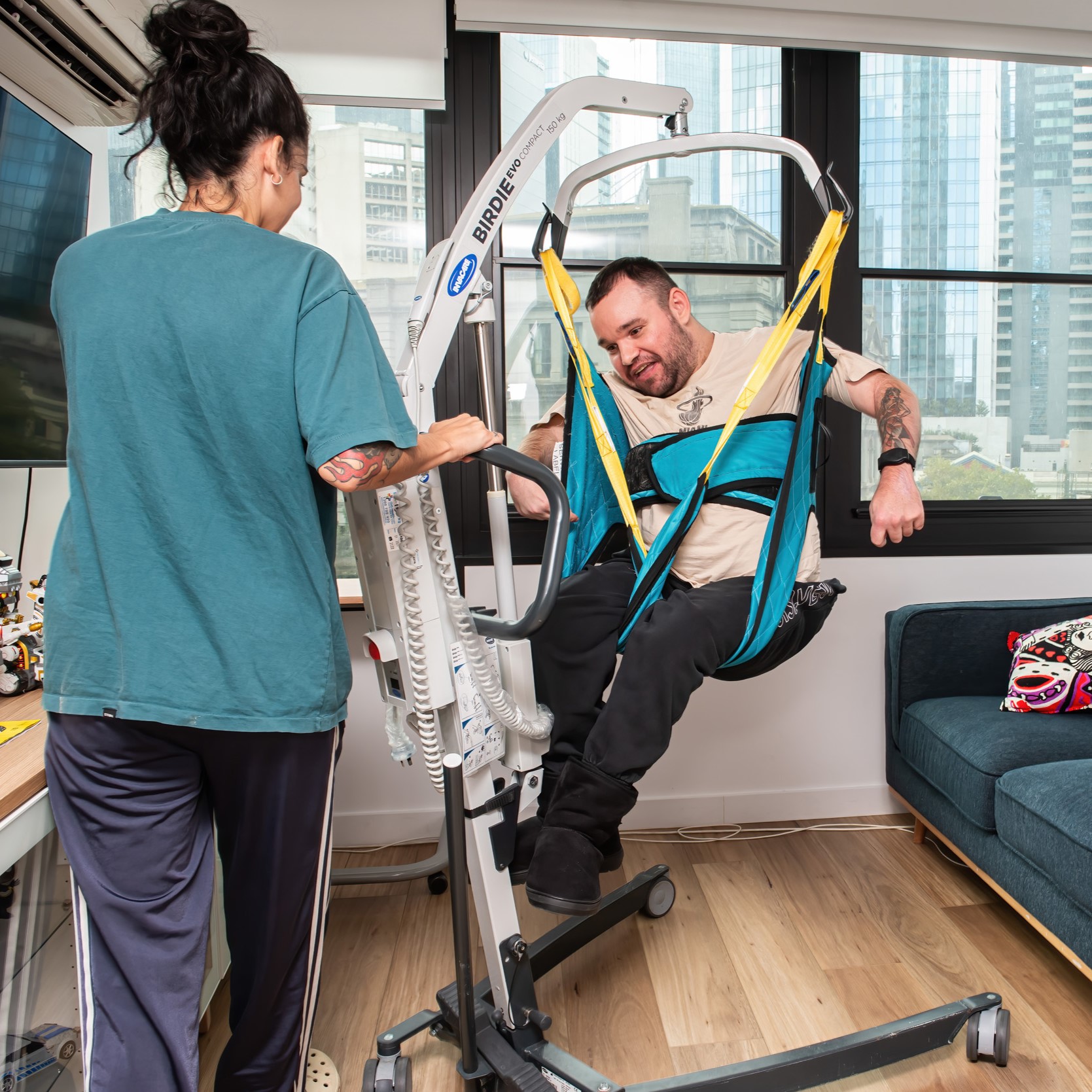 Jono is in air strapped into the hoist. He is wearing a white tshirt and black pants. His hoist sling is green. His support worker is on the left. She is wearing an aqua coloured tshirt and blue track pants. Windows in the background