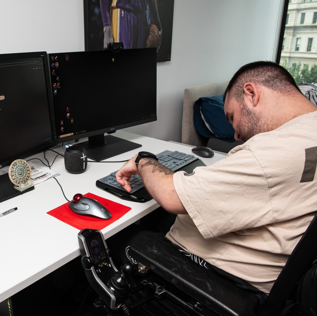 Jonathan, a non-verbal man with cerebral palsy, uses a trackball mouse and adaptive keyboard at his computer workstation. Seated in his powered wheelchair, he types using one finger while working at a desk with dual monitors in his apartment.